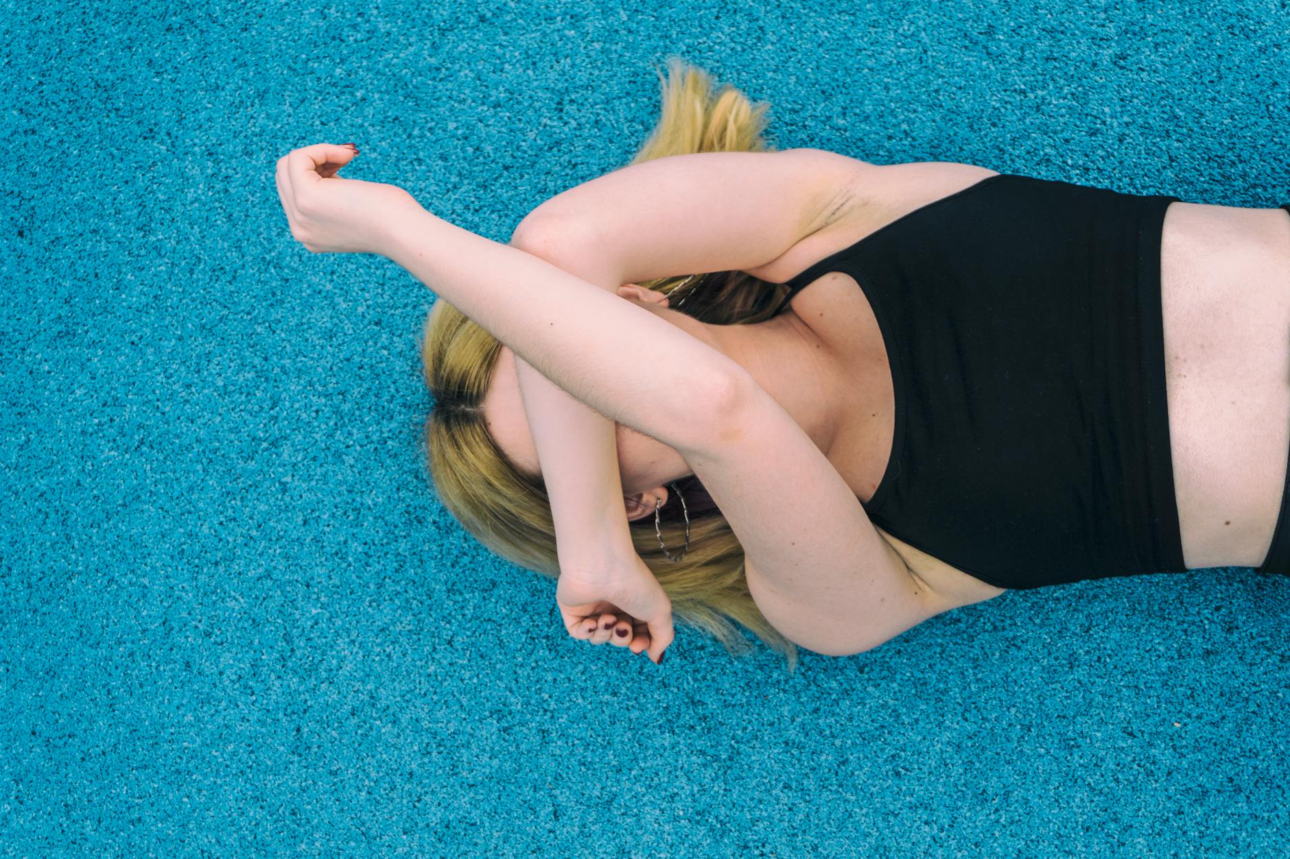 woman wearing black sports brassiered lying on teal surface while covering her face with her arms